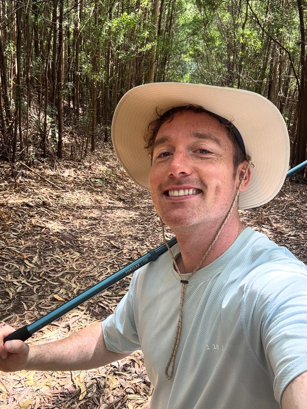 Christopher Adams in a wide-brimmed hat with a walking pole, grinning in a pine wood