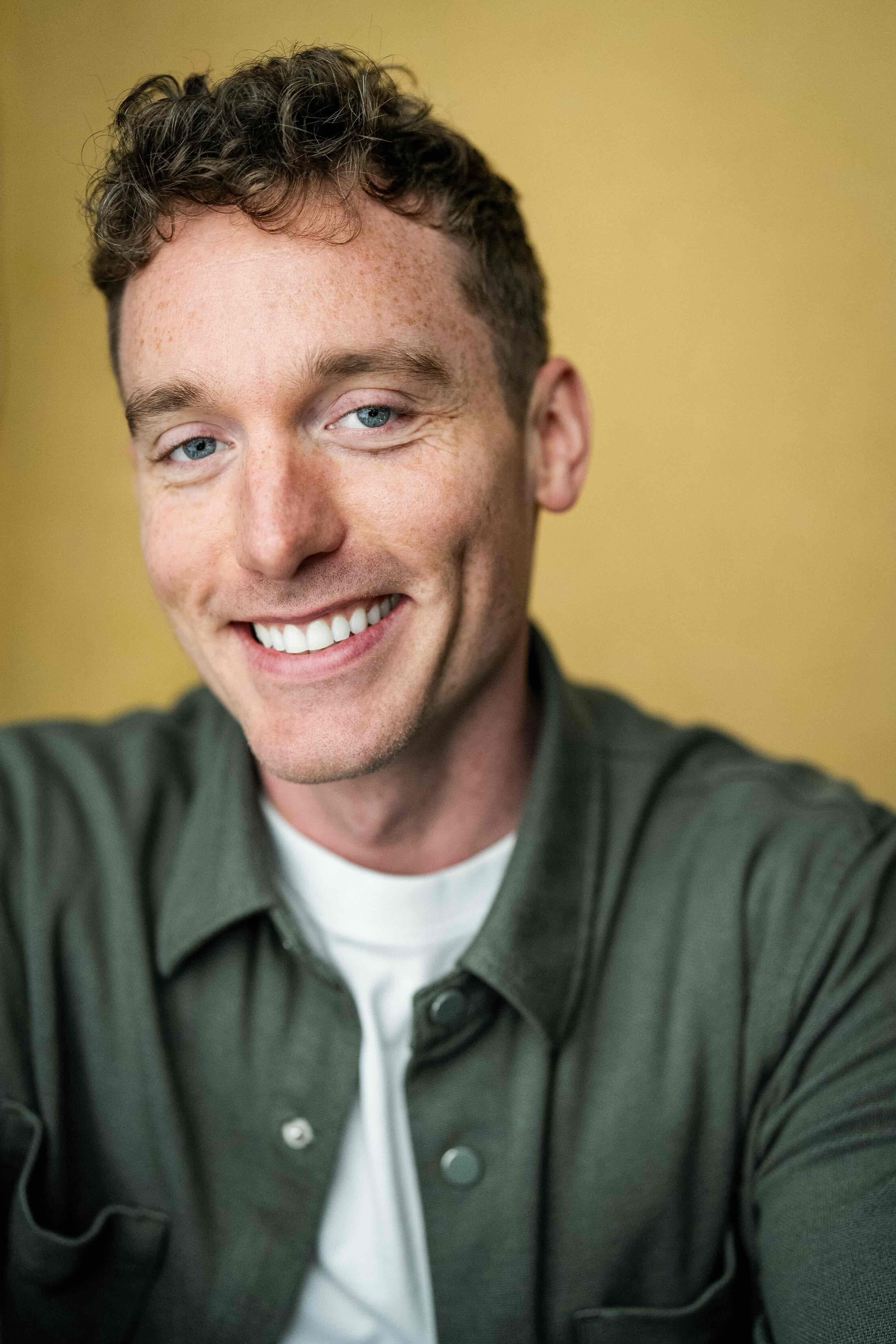 Christopher Adams headshot, smiling, green overshirt on a mustard backdrop