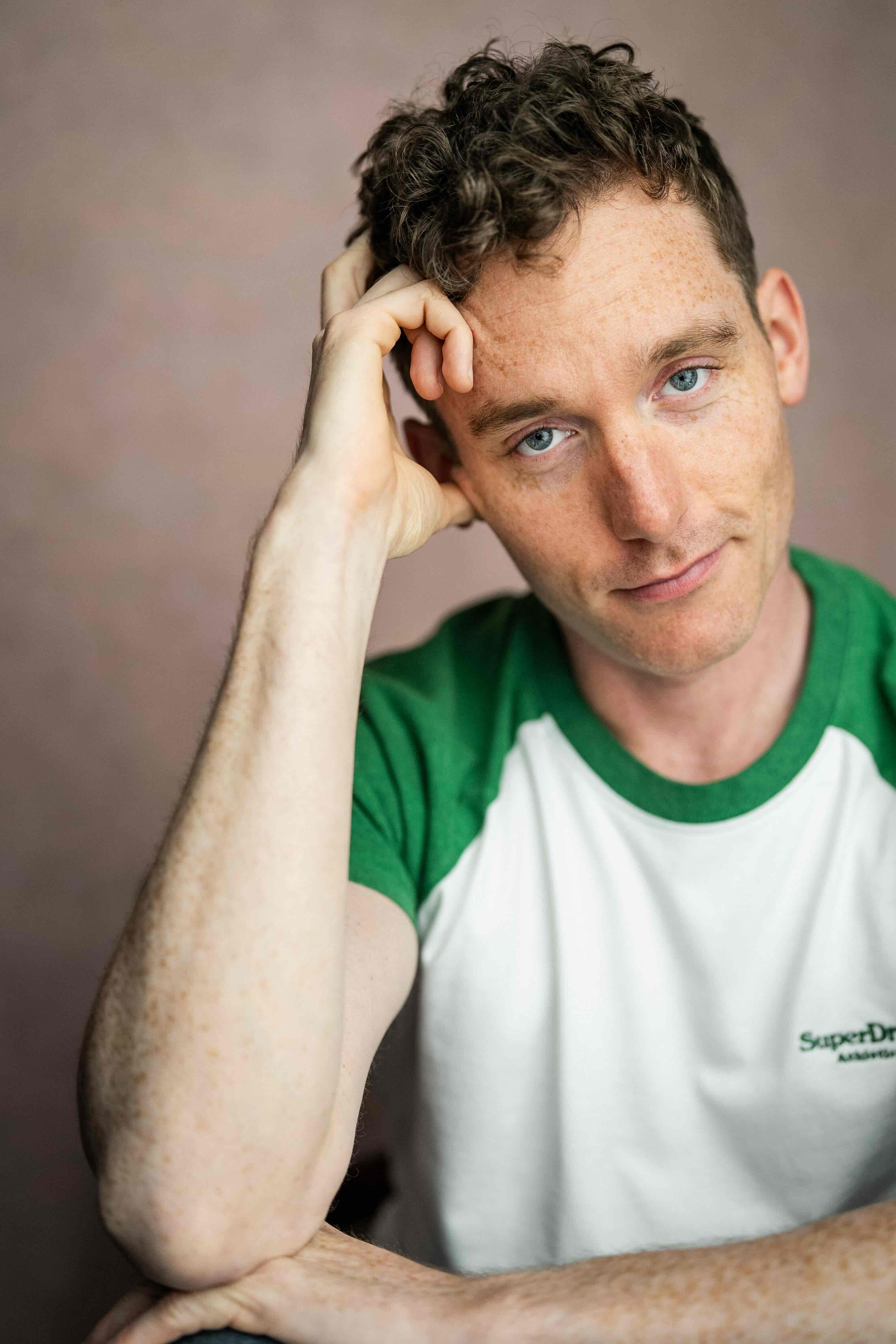 Christopher Adams headshot, head resting on one hand, green and white t-shirt on a dusty pink backdrop