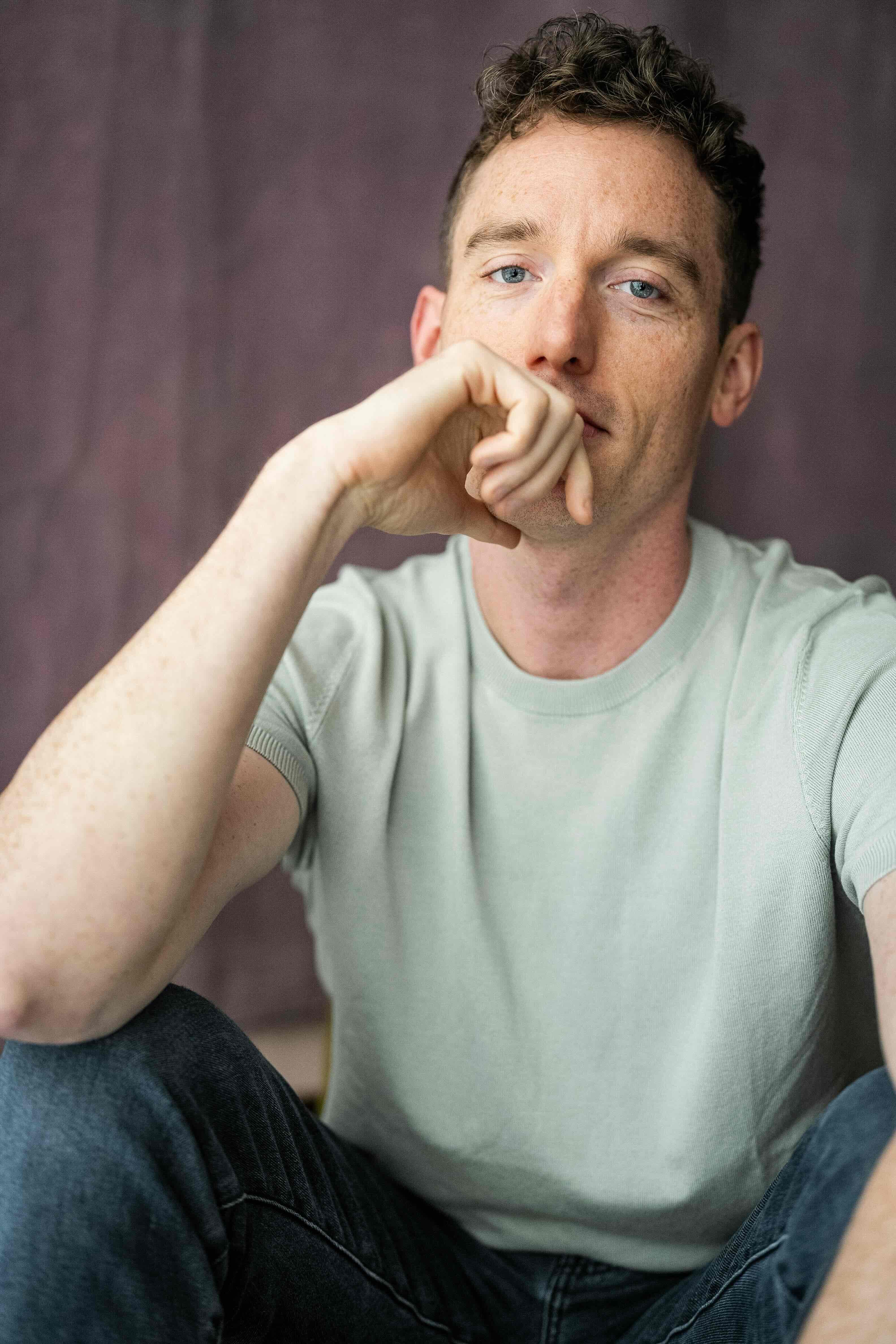 Christopher Adams headshot, seated with a hand at his chin, pale sage t-shirt on a mauve backdrop
