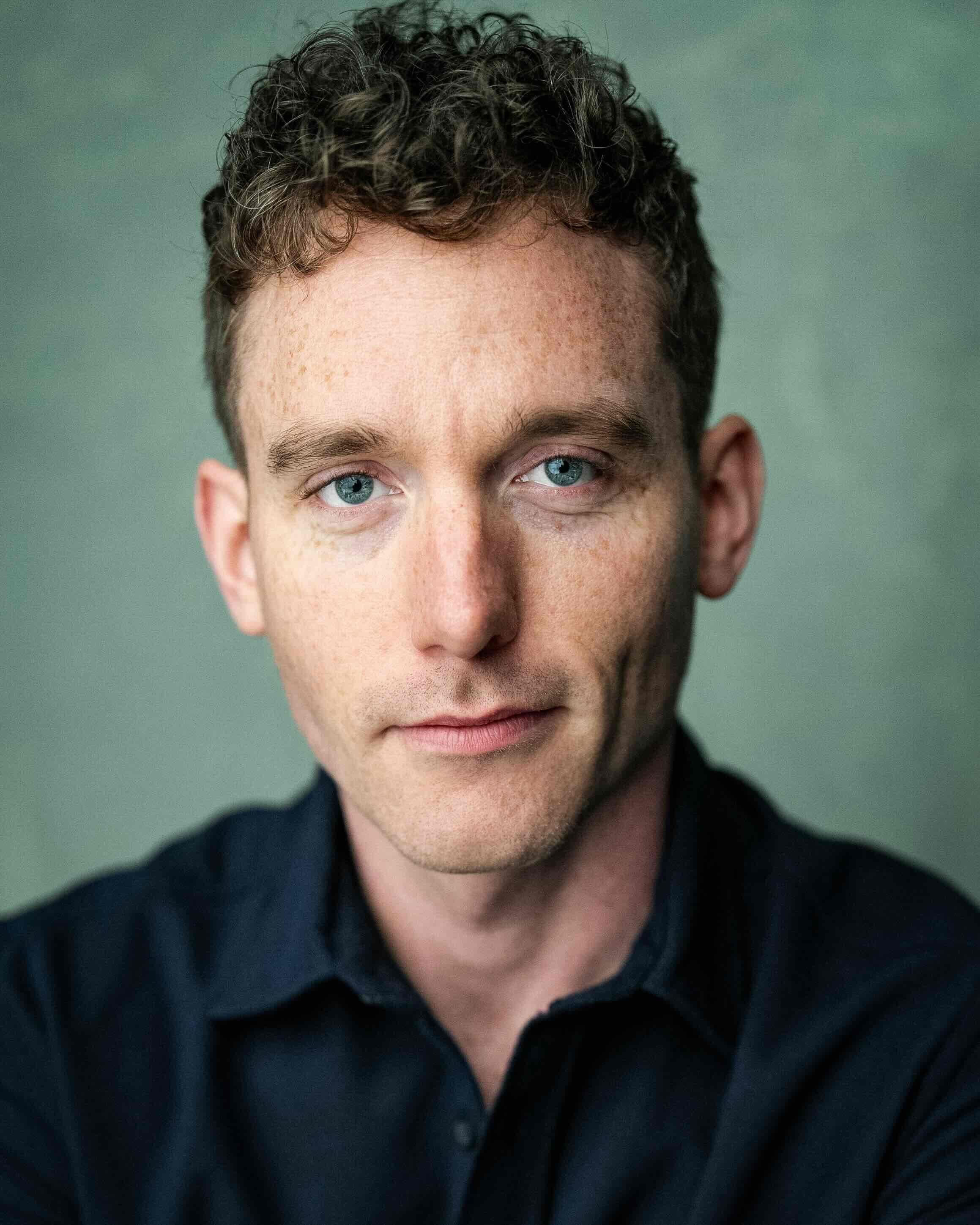 Christopher Adams headshot, close and straight to camera, navy shirt on a grey-green backdrop