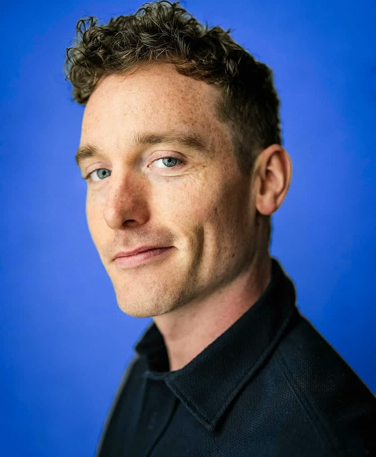 Christopher Adams, headshot, head tilted, dark shirt on a royal blue backdrop